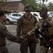 Three military personnel are engaged in a handshake greeting in what appears to be a tropical outdoor setting. Two individuals wearing U.S. Marine Corps MARPAT camouflage uniforms are interacting with a third individual in multicam/OCP pattern uniform with a tactical vest and cap bearing unit insignia. The setting suggests a joint military exercise or liaison meeting, likely in a Central American or Caribbean location based on the tropical vegetation and vehicle markings visible in the background.