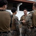 A group of military personnel in MARPAT (Marine pattern) camouflage uniforms are gathered in an outdoor briefing or discussion. One individual facing the camera appears to be speaking or instructing the others, gesturing with his hands. The setting appears to be a military installation or training area.