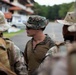 A group of military personnel are gathered outdoors in what appears to be a tropical or subtropical environment. The central figure wears USMC MARPAT digital camouflage with a boonie cover and sunglasses, while surrounding personnel wear OCP (Operational Camouflage Pattern) uniforms with tactical backpacks. The setting suggests a multinational or joint military exercise or operation.