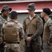 Four military personnel are engaged in a group discussion outdoors near a building with a red metal roof. Three individuals are wearing MARPAT (Marine digital camouflage) uniforms, while one person facing away is wearing a tan/multicam uniform with a plate carrier vest. The interaction appears to be a briefing or informal coordination between service members.