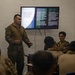 A military briefing or training session is taking place in a room where a presenter in U.S. Marine Corps camouflage uniform is delivering a presentation to a mixed audience of military personnel. The slide on the display screen covers the topic 'Communication With Maneuver / Comunicación con maniobra,' suggesting a bilingual (English/Spanish) training environment. The audience includes personnel in various military uniforms, indicating a multinational or joint training exercise.