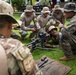 A group of military personnel in mixed camouflage uniforms are gathered on a grass area examining a weapon system mounted on a bipod. One soldier is positioned prone or low behind the weapon while others crouch around observing, taking notes, or using mobile devices. The scene appears to be an instructional or familiarization training session with the weapon system.
