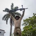 A soldier in full camouflage uniform holds a light machine gun overhead with both arms extended, standing on a grass area. The setting appears to be a tropical military installation, evidenced by palm trees, low-rise buildings with red-tiled roofs, and lush vegetation. The posture suggests a physical training exercise or a weapon-overhead drill.