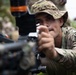 A soldier in multicam camouflage uniform and boonie hat is closely examining or adjusting a weapon system equipped with an optical sight. A patch on his right shoulder appears to show a unit insignia along with a blood type indicator reading 'O POS'. Other personnel in camouflage uniforms are partially visible in the background.