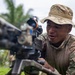 A soldier in multicam/OCP camouflage uniform and boonie hat is examining or operating a crew-served weapon, likely an M2 .50 caliber machine gun mounted on a tripod. A patch is visible on his left sleeve, and another individual in MARPAT (Marine) camouflage is partially visible to the left. The setting appears to be an outdoor training environment with tropical vegetation in the background.
