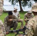 A group of military personnel in various camouflage uniforms are gathered outdoors near a waterfront, examining or training with a crew-served weapon mounted on a bipod. The setting appears to be a grassy area under trees with a body of water and a vessel visible in the background. This appears to be a joint military training exercise or weapons familiarization session.