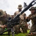 Three military personnel in camouflage uniforms are gathered around a crew-served weapon mounted on a tripod, appearing to conduct a training exercise. One soldier is actively handling the weapon while the others observe closely. The setting appears to be an outdoor grassy area near a building with palm trees visible in the background, suggesting a tropical location.