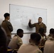 A military classroom instruction session is taking place, with two instructors in MARPAT (Marine) camouflage uniforms presenting to a group of students in tan/MultiCam uniforms. One instructor is gesturing toward a whiteboard that displays a tactical diagram labeled 'MORT' with arrows and movement indicators. The setting appears to be a formal military training environment.