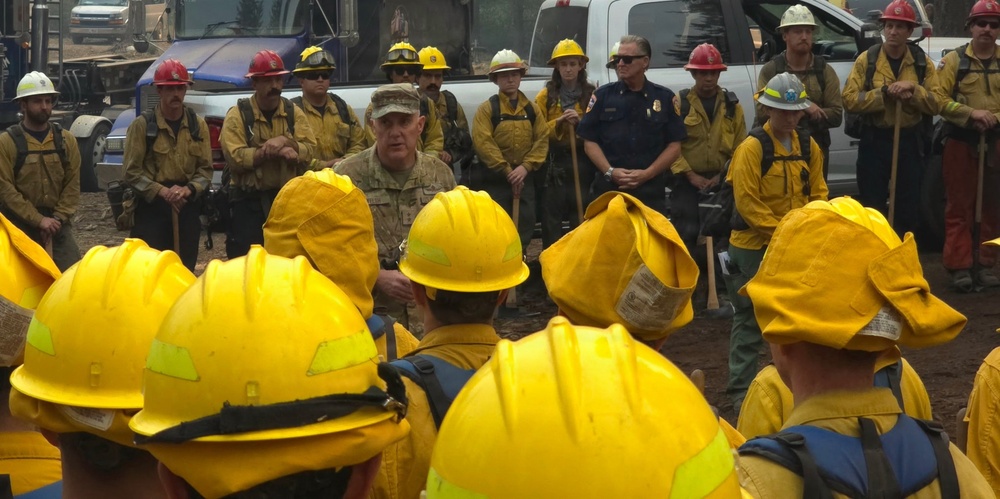 A large group of wildland firefighters in yellow Nomex gear and hard hats are gathered in a semicircle for what appears to be a briefing or debriefing session outdoors. A person in military camouflage uniform appears to be addressing the group, alongside individuals in blue fire department uniforms and standard firefighting attire. The setting appears to be a staging area or incident command post, with trucks and equipment visible in the background.