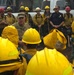 A large group of wildland firefighters in yellow Nomex gear and hard hats are gathered in a semicircle for what appears to be a briefing or debriefing session outdoors. A person in military camouflage uniform appears to be addressing the group, alongside individuals in blue fire department uniforms and standard firefighting attire. The setting appears to be a staging area or incident command post, with trucks and equipment visible in the background.