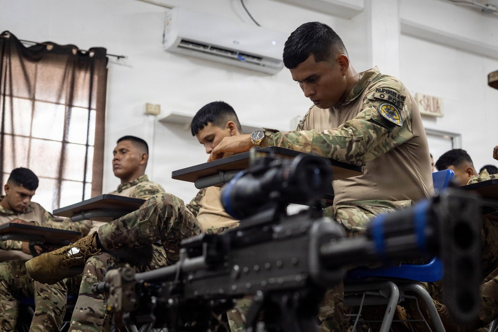 Military or law enforcement personnel in camouflage uniforms are seated at student desks in a classroom setting, appearing to be taking a written examination or attending a training course. A weapon with an optical scope is visible in the foreground, suggesting this is a weapons or tactical training class. A wall-mounted air conditioning unit and a 'SALIDA' (exit) sign are visible, indicating a Spanish-speaking country's facility.