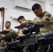 Military or law enforcement personnel in camouflage uniforms are seated at student desks in a classroom setting, appearing to be taking a written examination or attending a training course. A weapon with an optical scope is visible in the foreground, suggesting this is a weapons or tactical training class. A wall-mounted air conditioning unit and a 'SALIDA' (exit) sign are visible, indicating a Spanish-speaking country's facility.