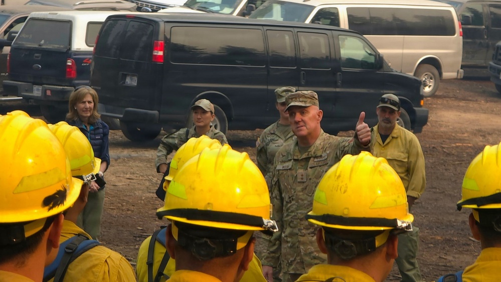 A military officer in camouflage uniform gives a thumbs-up while addressing a group of wildland firefighters wearing yellow hard hats and fire-resistant gear. The briefing or morale visit appears to take place at a staging area or base camp, with multiple vehicles parked in the background. Several other individuals, including civilians and additional military personnel, are present in the background.