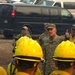 A military officer in camouflage uniform gives a thumbs-up while addressing a group of wildland firefighters wearing yellow hard hats and fire-resistant gear. The briefing or morale visit appears to take place at a staging area or base camp, with multiple vehicles parked in the background. Several other individuals, including civilians and additional military personnel, are present in the background.