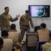 Two U.S. Marines in MARPAT camouflage uniforms are conducting a bilingual briefing titled 'Leaders Recon / líderes reconocen' to a group of seated military personnel wearing mixed uniforms. The presentation is displayed on a flat-screen monitor mounted on a white wall. The setting appears to be a classroom or briefing room in a joint or multinational training environment.
