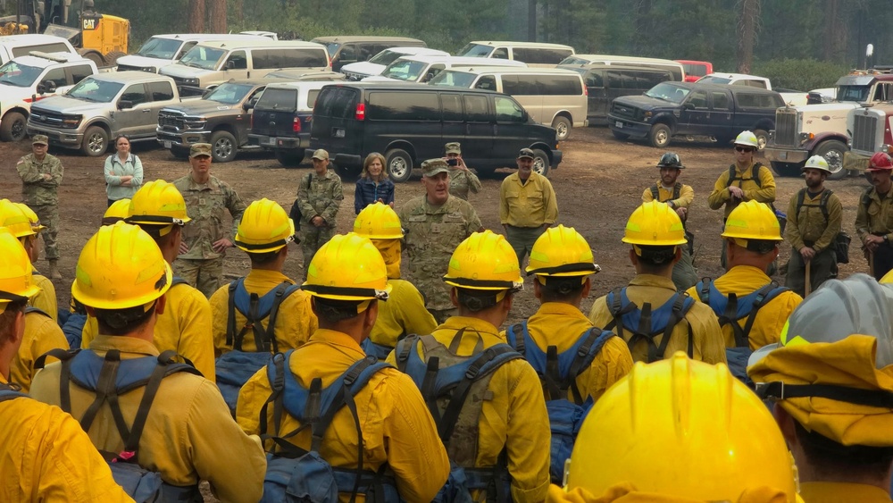 A large group of wildland firefighters wearing yellow hard hats and Nomex fire-resistant gear are assembled in a semicircle at a staging area, receiving a briefing from military and civilian personnel. The setting appears to be a forest clearing used as an incident command or base camp, with numerous vehicles including vans, trucks, and heavy equipment visible in the background. The scene suggests a coordinated wildfire response operation involving both military and civilian firefighting resources.
