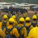 A large group of wildland firefighters wearing yellow hard hats and Nomex fire-resistant gear are assembled in a semicircle at a staging area, receiving a briefing from military and civilian personnel. The setting appears to be a forest clearing used as an incident command or base camp, with numerous vehicles including vans, trucks, and heavy equipment visible in the background. The scene suggests a coordinated wildfire response operation involving both military and civilian firefighting resources.