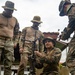 Military personnel in camouflage uniforms are gathered outdoors on a grassy area, appearing to assemble or demonstrate a weapon system mounted on a tripod. One soldier is kneeling and handling the weapon while others observe. The group includes personnel wearing different camouflage patterns, suggesting a joint or multinational training exercise.