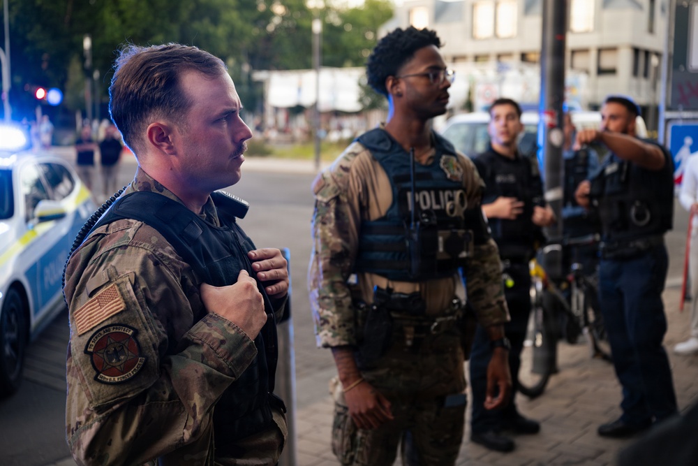 The image shows a group of law enforcement or military personnel standing on a sidewalk in what appears to be an urban setting. The individuals are wearing camouflage uniforms and tactical vests, with at least one vest visibly labeled 'POLICIO'. Emergency vehicle lights are visible in the background, suggesting an active incident or operation.