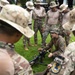 A group of military personnel in camouflage uniforms are gathered outdoors on a grass area during what appears to be a weapons familiarization or training session. One individual in Marine-pattern camouflage is kneeling and demonstrating a machine gun mounted on a bipod to the surrounding group. The setting appears to be a military training environment, possibly a multinational exercise given the varied uniform styles.