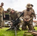 Military personnel in camouflage uniforms are engaged in a weapons training or familiarization exercise outdoors on a grass area. Two soldiers are kneeling and handling what appears to be a tripod-mounted grenade launcher or heavy weapon system. The setting appears to be a military installation or compound, with additional personnel observing.