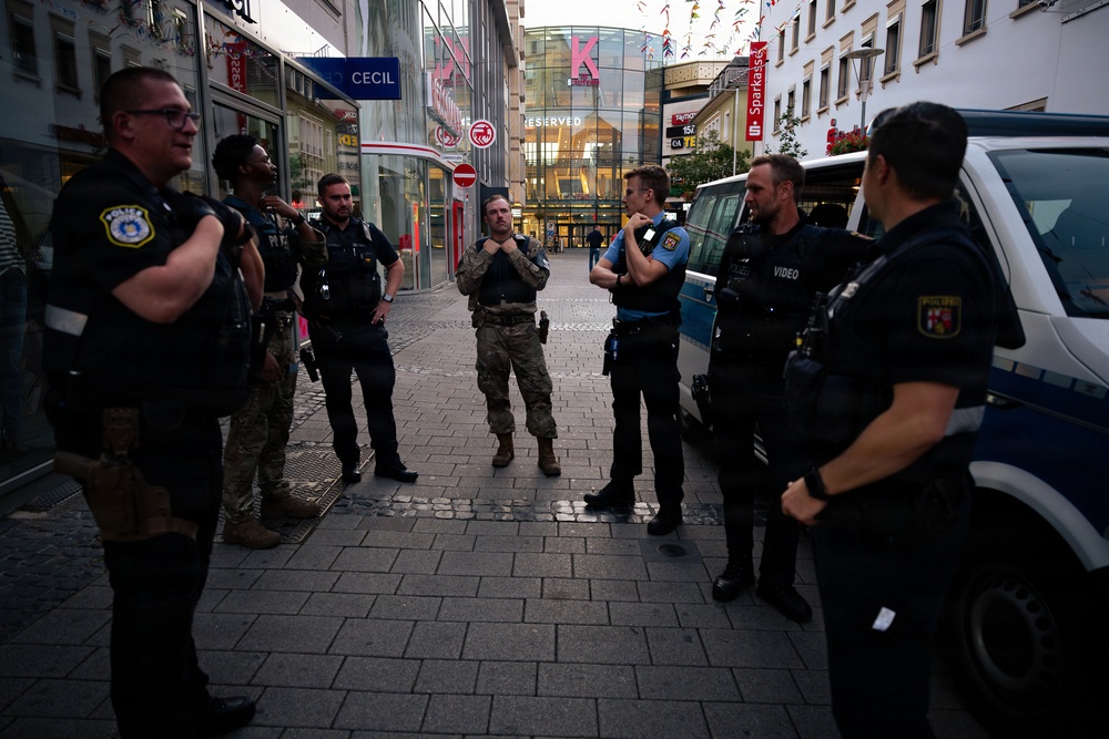 A group of law enforcement personnel are gathered in a pedestrian shopping street, appearing to coordinate or debrief. The group includes individuals in German police uniforms (Polizei), a person in military-style camouflage, and at least one officer with a 'Police' patch suggesting possible US military police presence. Visible storefronts and signage indicate a German city center environment.