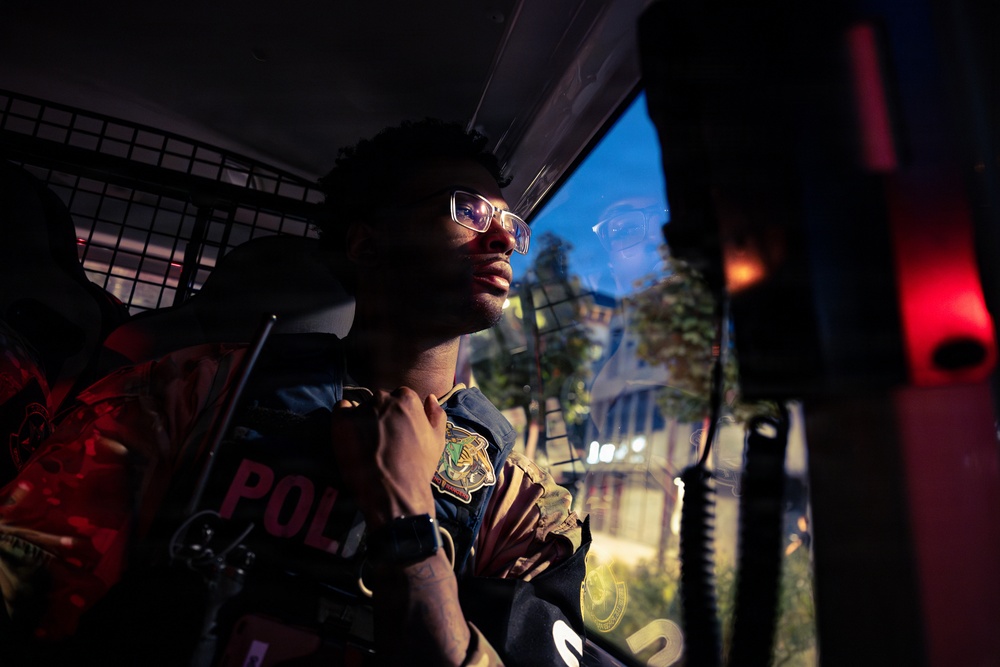 A person wearing glasses and what appears to be a police or security vest is seated inside a vehicle, looking out through a window. The scene is dramatically lit with red and blue tones, suggesting a nighttime law enforcement context. A metal security partition/cage is visible in the background of the vehicle interior.