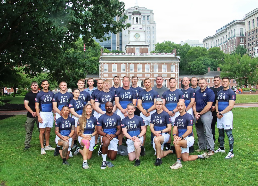 A large group of approximately 25 individuals poses for a team photo on a grass lawn wearing 'Team USA' blue and white athletic uniforms. They are positioned in front of what appears to be Independence Hall in Philadelphia, Pennsylvania. The group is arranged in three rows with some members kneeling in front and others standing in the back.
