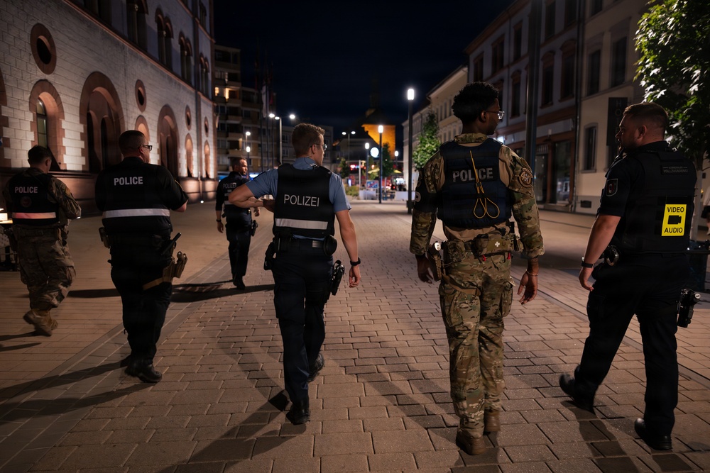 A joint patrol of American military police and German police (Polizei) walking together through a city street at night. The personnel are wearing a mix of U.S. military camouflage uniforms with 'POLICE' vests and German police uniforms labeled 'POLIZEI'. This appears to be a cooperative law enforcement patrol, likely near a U.S. military installation in Germany.