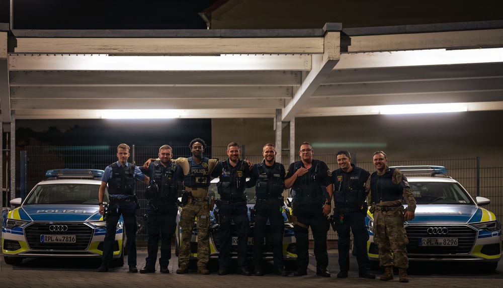 A group of eight men pose together at night in what appears to be a German police facility or parking area. The group includes both German police officers in standard blue uniforms marked 'POLIZEI' and individuals in military-style tactical/camouflage uniforms, suggesting a joint law enforcement or military-police cooperation event. Two Audi police vehicles with German license plates are visible flanking the group.