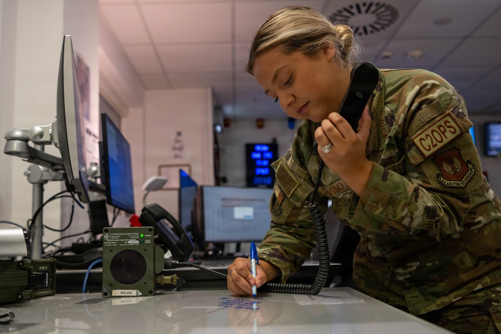 A female U.S. Air Force service member in OCP (Operational Camouflage Pattern) uniform is working at a command and control operations desk. She is simultaneously talking on a corded telephone handset while writing notes on a surface, suggesting active coordination duties. The setting appears to be a military operations center with multiple monitors and communications equipment visible.