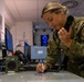 A female U.S. Air Force service member in OCP (Operational Camouflage Pattern) uniform is working at a command and control operations desk. She is simultaneously talking on a corded telephone handset while writing notes on a surface, suggesting active coordination duties. The setting appears to be a military operations center with multiple monitors and communications equipment visible.