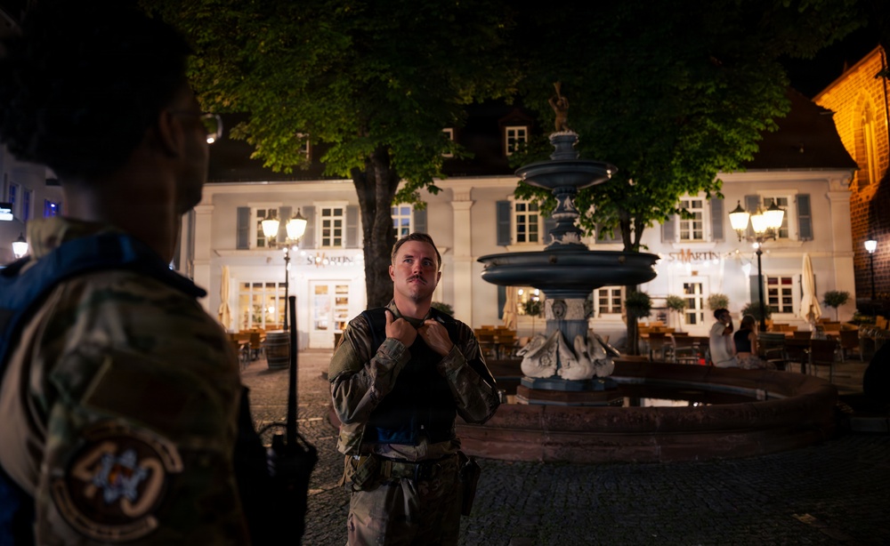 Two military personnel in camouflage uniforms are conversing in a European town square at night. A decorative multi-tiered fountain is visible in the background, along with an illuminated building bearing the sign 'St. Martin'. The setting appears to be a German or Central European town center.