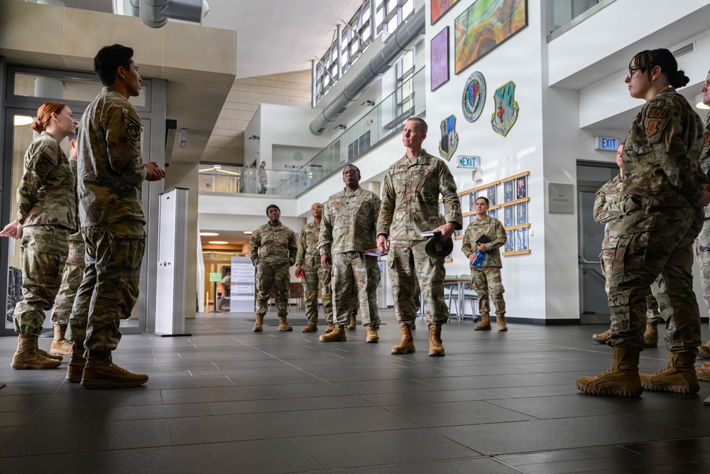 A group of military personnel in OCP (Operational Camouflage Pattern) uniforms are gathered in what appears to be the lobby or atrium of a military facility. A senior officer holding a cover (hat) and documents appears to be addressing or briefing the assembled group. The setting includes visible unit patches and insignia on the walls, consistent with a US Air Force installation.