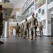 A group of military personnel in OCP (Operational Camouflage Pattern) uniforms are gathered in what appears to be the lobby or atrium of a military facility. A senior officer holding a cover (hat) and documents appears to be addressing or briefing the assembled group. The setting includes visible unit patches and insignia on the walls, consistent with a US Air Force installation.
