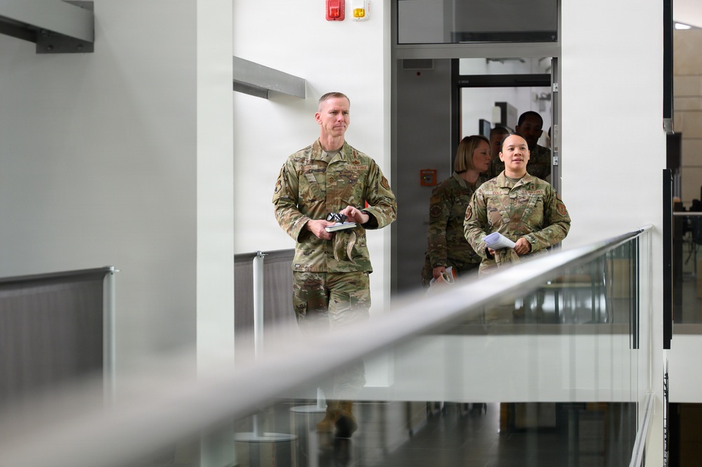 Two military personnel in OCP (Operational Camouflage Pattern) uniforms are walking through what appears to be a modern military or government building interior. A taller male service member leads, holding a small device or notebook, while a shorter female service member follows holding papers. Additional personnel in uniform and civilian clothing are visible in the background.