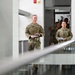 Two military personnel in OCP (Operational Camouflage Pattern) uniforms are walking through what appears to be a modern military or government building interior. A taller male service member leads, holding a small device or notebook, while a shorter female service member follows holding papers. Additional personnel in uniform and civilian clothing are visible in the background.