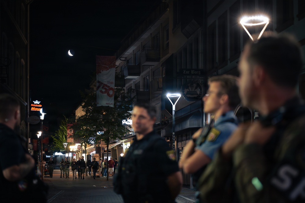 A nighttime street scene showing multiple uniformed officers standing in a pedestrian zone, appearing to monitor or secure the area. The street is lined with commercial buildings, illuminated signage, and modern street lamps. A crescent moon is visible in the dark sky above the urban corridor.