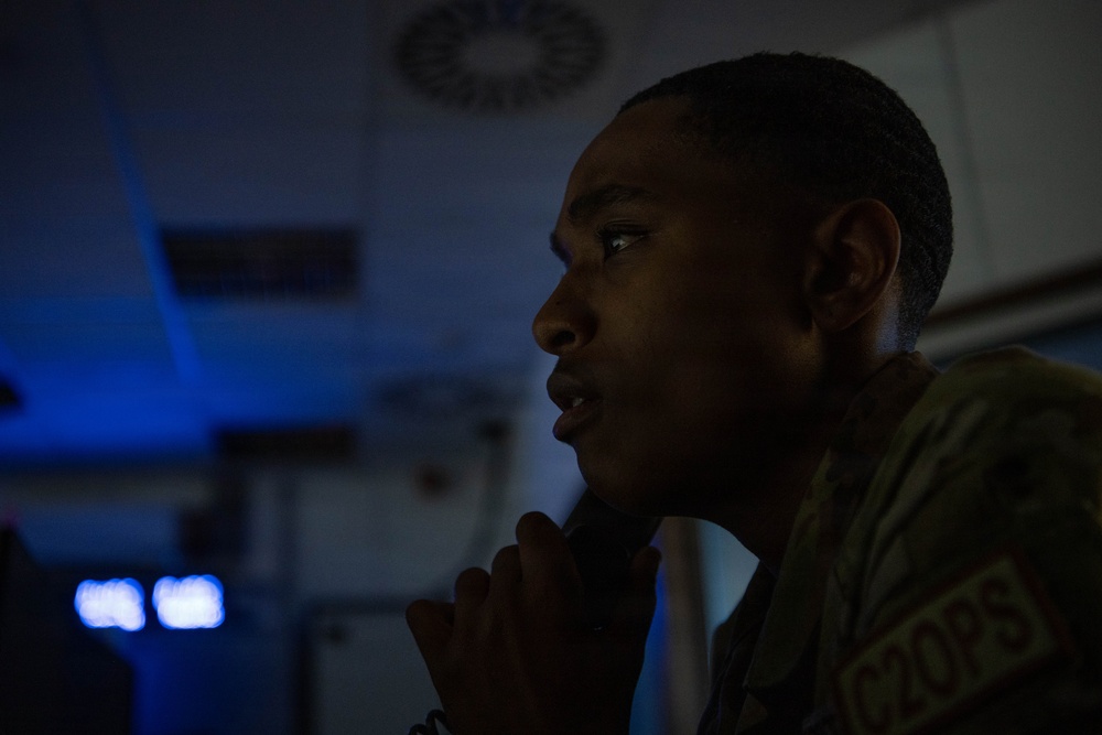 A young male individual wearing military or tactical clothing is photographed in profile in a dimly lit environment. The subject appears to be in a contemplative or attentive posture with hands clasped near the chin. Blue ambient lighting is visible in the background, suggesting an operational or command-type setting.