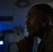 A young male individual wearing military or tactical clothing is photographed in profile in a dimly lit environment. The subject appears to be in a contemplative or attentive posture with hands clasped near the chin. Blue ambient lighting is visible in the background, suggesting an operational or command-type setting.