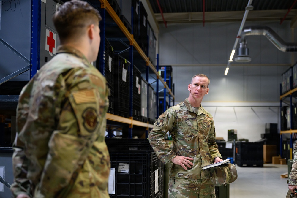 Two U.S. Air Force personnel in OCP (Operational Camouflage Pattern) uniforms are engaged in a conversation inside a military warehouse or supply depot. The individual facing the camera, identified by a name tape reading 'CLARK,' is holding a notebook and appears to be conducting or receiving a briefing. The setting contains shelving units stocked with military equipment and storage containers.