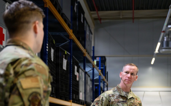 Two U.S. Air Force personnel in OCP (Operational Camouflage Pattern) uniforms are engaged in a conversation inside a military warehouse or supply depot. The individual facing the camera, identified by a name tape reading 'CLARK,' is holding a notebook and appears to be conducting or receiving a briefing. The setting contains shelving units stocked with military equipment and storage containers.
