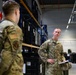Two U.S. Air Force personnel in OCP (Operational Camouflage Pattern) uniforms are engaged in a conversation inside a military warehouse or supply depot. The individual facing the camera, identified by a name tape reading 'CLARK,' is holding a notebook and appears to be conducting or receiving a briefing. The setting contains shelving units stocked with military equipment and storage containers.