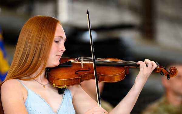 A young woman with long red hair is playing a violin at what appears to be a military ceremony or event. She is wearing a light blue sundress and is focused on her performance. In the background, military personnel in camouflage uniforms are visible, along with what appears to be a blue military flag.