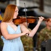 A young woman with long red hair is playing a violin at what appears to be a military ceremony or event. She is wearing a light blue sundress and is focused on her performance. In the background, military personnel in camouflage uniforms are visible, along with what appears to be a blue military flag.