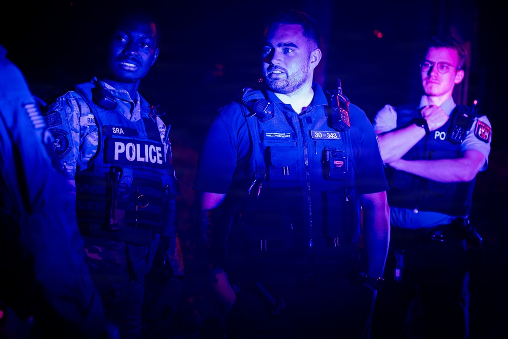 Three law enforcement officers are standing together in a low-light environment illuminated by blue lighting. The officers appear to represent different agencies, with visible insignia including 'POLICE' on a tactical vest, 'SRA' rank designation, and what appears to be German 'Vollzugsdienst' (enforcement service) markings with badge number 30-343. The scene suggests a joint operation or coordinated law enforcement activity.