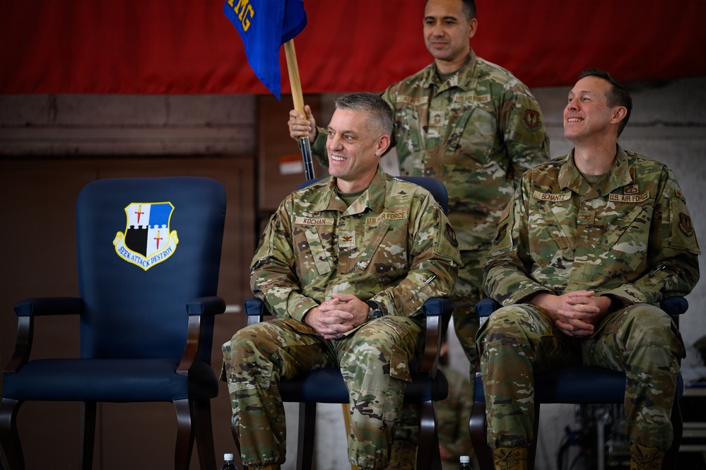 Three U.S. Air Force personnel in OCP (Operational Camouflage Pattern) uniforms are seated/standing at what appears to be a military ceremony, likely a change of command. Two officers identified by name tapes as 'KOCHAN' and 'SCHANTZ' are seated in the foreground smiling, while a third individual stands behind holding a blue guidon flag. A unit shield bearing the motto 'SEEK ATTACK DESTROY' is visible on a chair to the left.