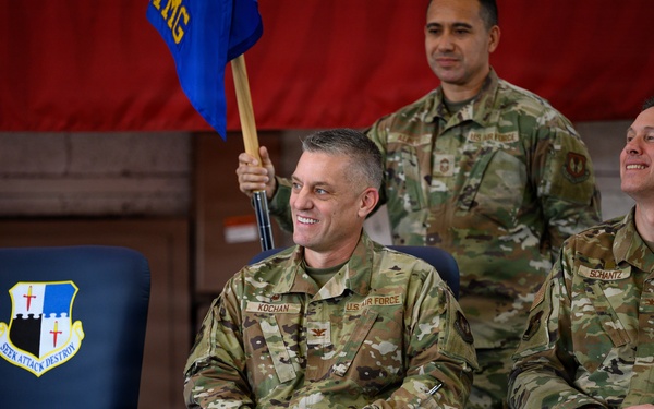Three U.S. Air Force personnel in OCP (Operational Camouflage Pattern) uniforms are seated/standing at what appears to be a military ceremony, likely a change of command. Two officers identified by name tapes as 'KOCHAN' and 'SCHANTZ' are seated in the foreground smiling, while a third individual stands behind holding a blue guidon flag. A unit shield bearing the motto 'SEEK ATTACK DESTROY' is visible on a chair to the left.
