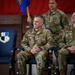 Three U.S. Air Force personnel in OCP (Operational Camouflage Pattern) uniforms are seated/standing at what appears to be a military ceremony, likely a change of command. Two officers identified by name tapes as 'KOCHAN' and 'SCHANTZ' are seated in the foreground smiling, while a third individual stands behind holding a blue guidon flag. A unit shield bearing the motto 'SEEK ATTACK DESTROY' is visible on a chair to the left.