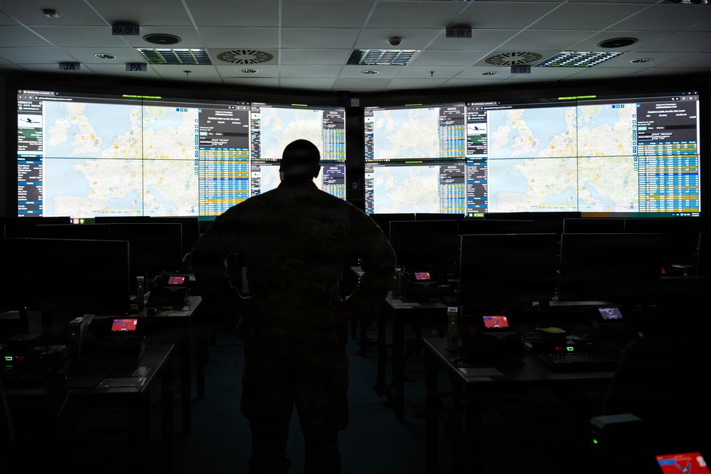A silhouetted individual in military camouflage uniform stands before a large multi-screen video wall displaying geographic maps and data tables in a darkened operations or command center. The room contains multiple workstations with monitors, keyboards, and equipment arranged in rows. The large display wall appears to show air traffic or operational tracking information over European geography.